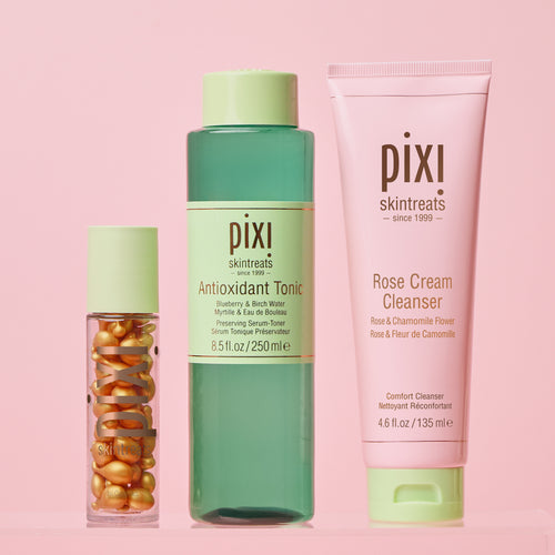 Three Pixi Skintreats skincare products arranged on a soft pink background: a small clear bottle with gold capsules, a translucent green bottle labeled Antioxidant Tonic, and a pink tube labeled Rose Cream Cleanser. view 1