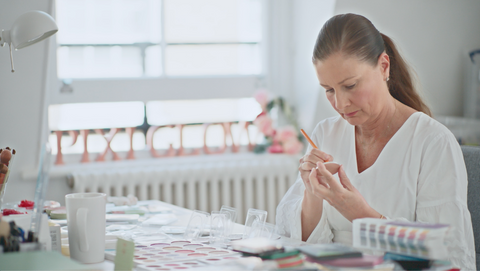A woman with her hair in a ponytail sits at a table, carefully examining a small clear object with a pencil. The room is bright and airy with a large window in the background, and the table is cluttered with various craft or sample materials.
