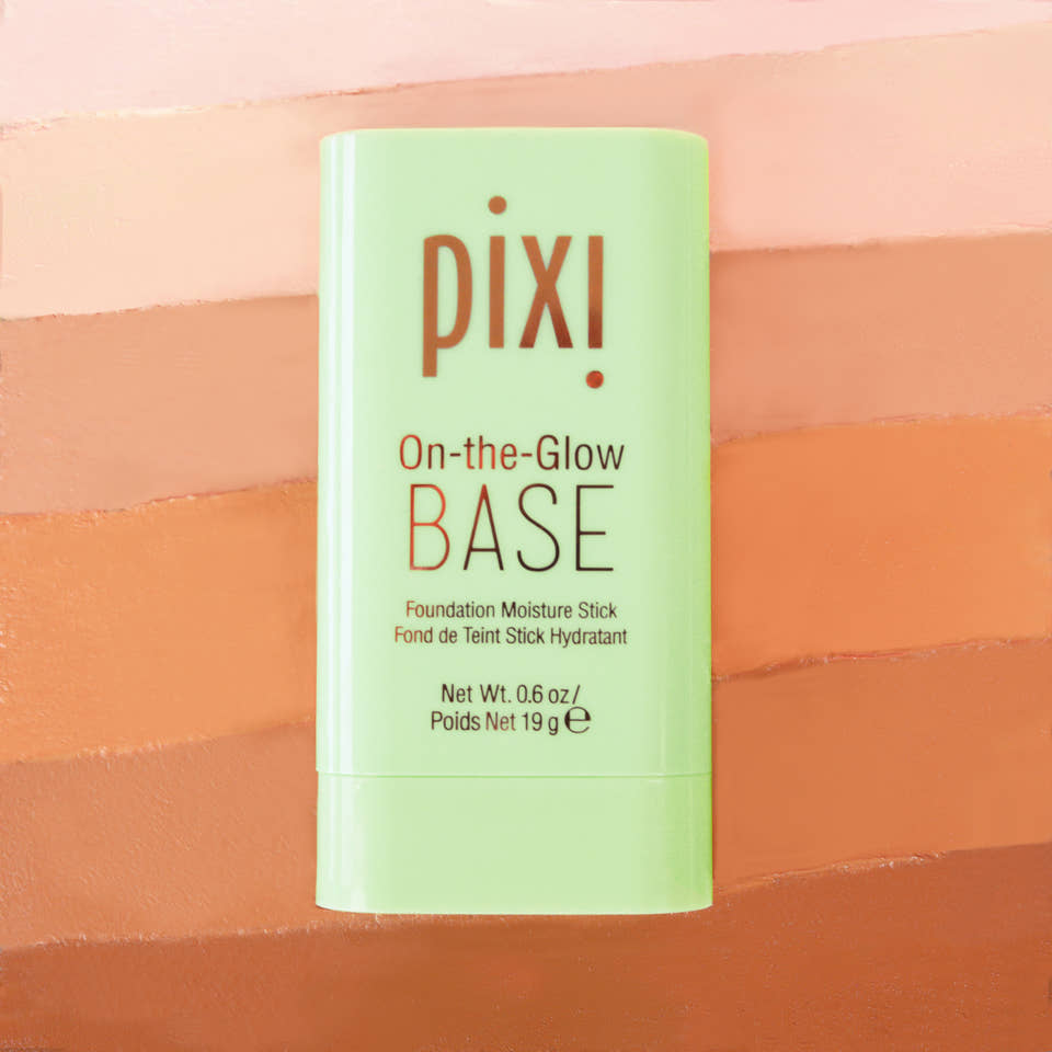 Pixi On-the-Glow Base Foundation Moisture Stick in a light green container, set against a background of horizontal peach and orange color swatches. view 1