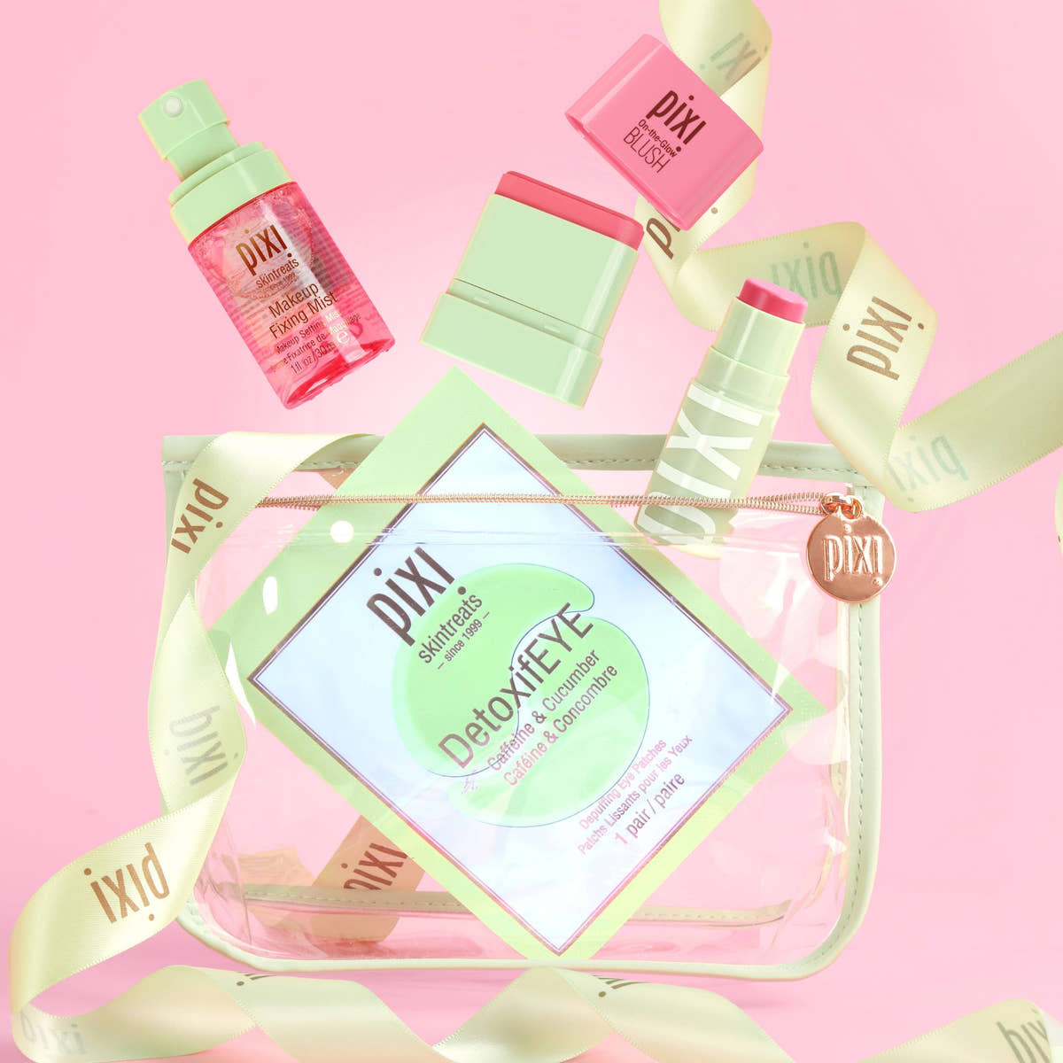 A Best of Pixi Beauty gift set arranged around a clear zip pouch on a pink background. The set includes a pink and mint detox eye cream tube, a pink and mint lip balm, a pink and mint lip product, a pink and mint cream product, and a clear pouch with a mint green border and Pixi branding.